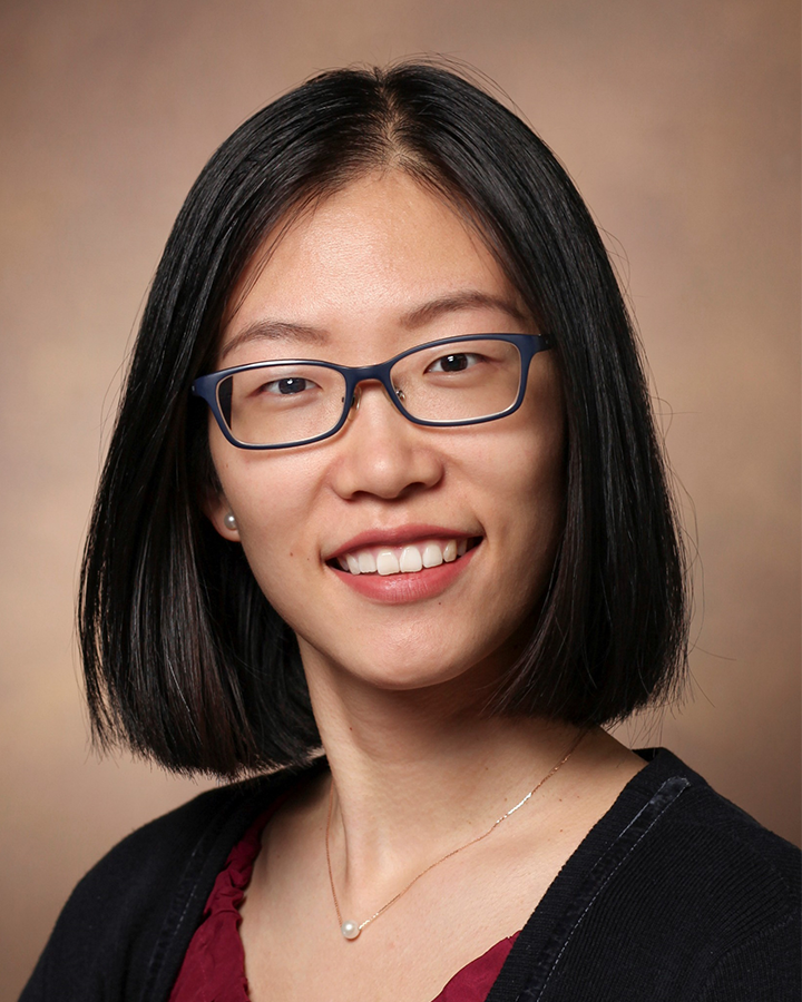 Sifang Kathy Zhao in front of a studio backdrop, wearing dark-framed glasses and a single pearl necklace