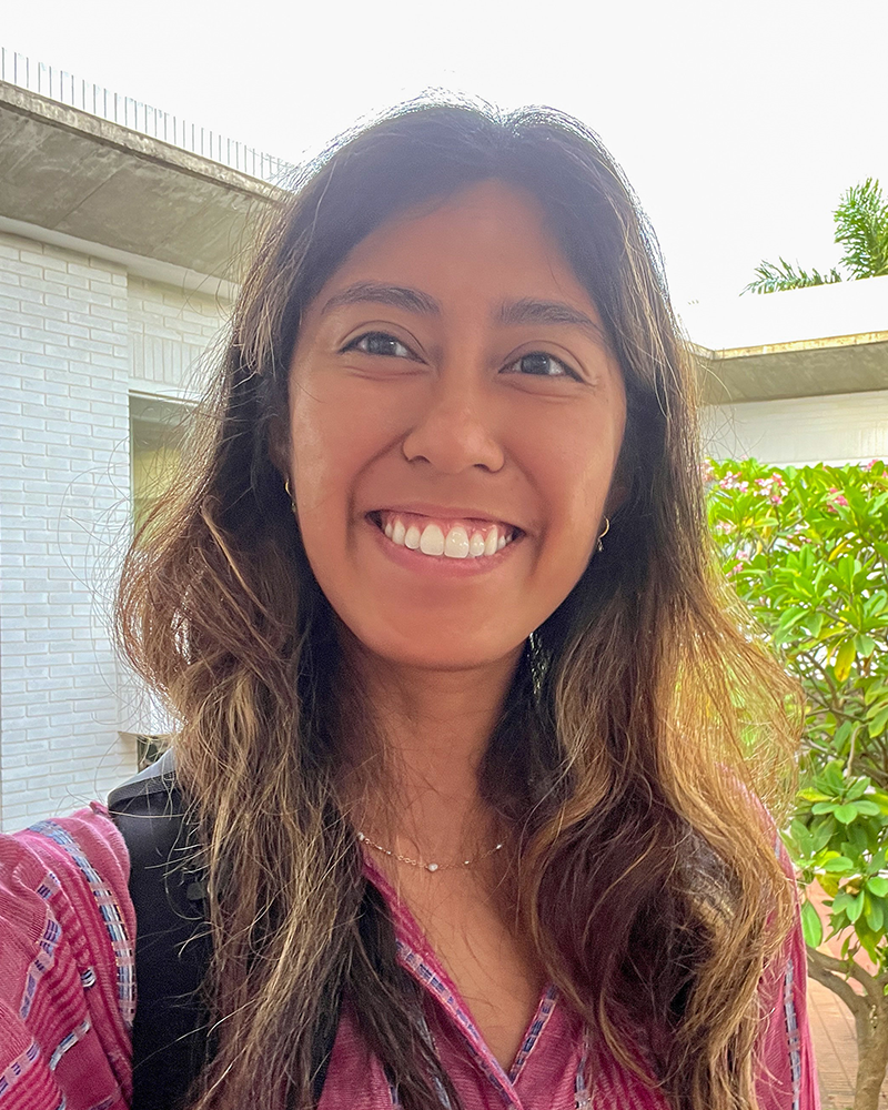 Frances Fernando smiling with a white brick building and a leafy green tree behind her