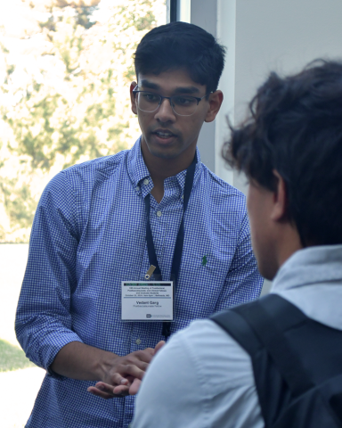 A fellow in a blue checked shirt speaks to another fellow in the foreground with their back to the camera