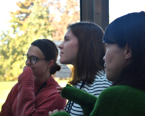 Three fellows look off to the left, out of frame. The leftmost in a red sweater, holds her hand to her mouth pensively. The rightmost fellow in the foreground gestures with a sleeve-covered hand.