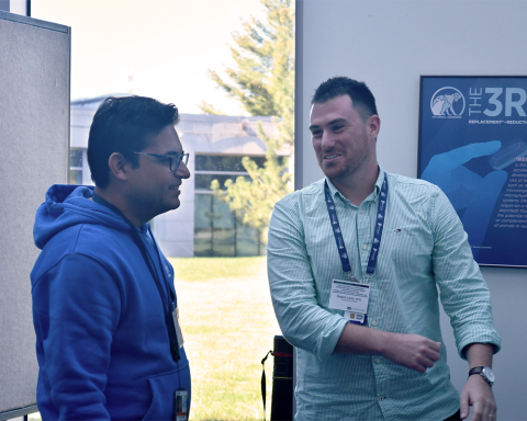 Two fellows chatting in front of a poster board and a large window, through which you can see grass, trees, and another building.