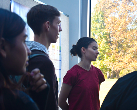 Three fellows look off to the right, out of frame. A large window behind them shows leafy trees in autumn colors.