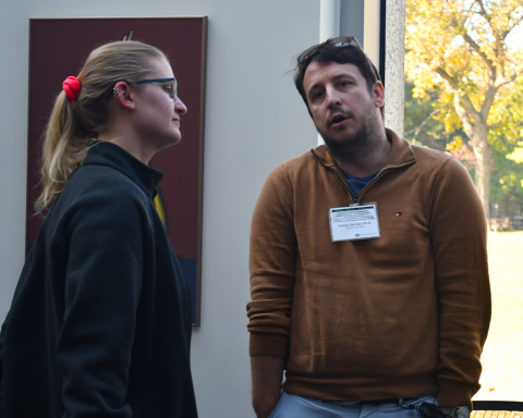 A blonde fellow with a ponytail talks to another with a brown sweatshirt