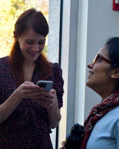 A fellow smiles widely as they look down at their phone. A second fellow, at right in profile, smiles while looking at something out of frame.