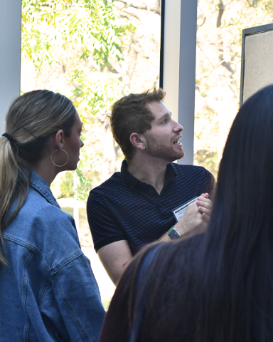 A fellow clasps their hands, mid-sentence, looking off to the right. Another fellow in a denim jacket listens.