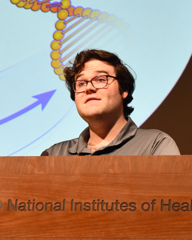 A fellow in glasses and a tan shirt stands behind a lectern. Behind them, an illustration of DNA is projected on a screen.