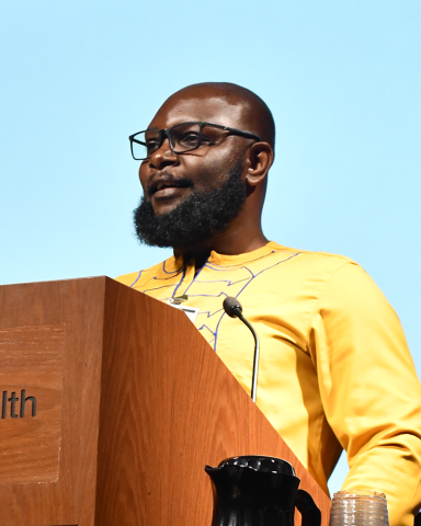 A bearded research fellow in a yellow shirt stands behind a lectern