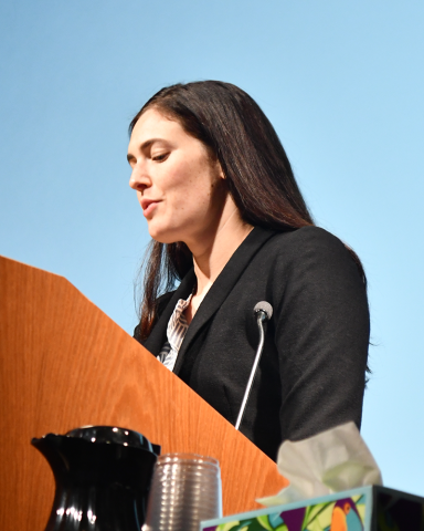 A woman in a black blazer looks down at her notes from a lectern